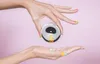 Two hands with colorful nail polish are shown against a pink background. One hand holds a pill dispenser, and the other hand holds two pills. Produktbild-Vorschau