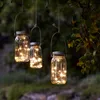 Three mason jar lanterns with fairy lights hanging from a tree branch at dusk. Produktbild-Vorschau