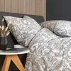 A close-up view of a bed with paisley patterned bedding and pillows, next to a small side table with a laptop and a vase of dried flowers. Produktbild-Vorschau