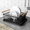 A black wire dish rack with plates, glasses, and utensils on a gray countertop in front of a white brick wall. Produktbild-Vorschau