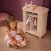 A young girl is playing with a doll next to a wooden doll wardrobe. Produktbild-Vorschau