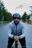 A man wearing a bicycle helmet and jacket is posing with his bicycle on a road. Náhled obrázku produktu