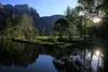 A serene river flows in the foreground, reflecting the trees and mountains in the background. The sun is setting or rising, casting a warm glow on the scene. Produktbild-Vorschau