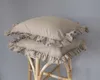 Two taupe pillows with ruffles are stacked on a light-colored wicker stool against a light gray background. Produktbild-Vorschau