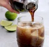A hand is pouring a dark liquid from a glass bottle into a glass filled with ice and a brown beverage. There are lime wedges and a whole lime in the background. Produktbild-Vorschau