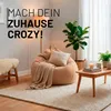 A cozy living room scene featuring a large beanbag chair, a small wooden coffee table with a candle and books, and plants. Voir les images du produit