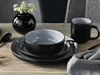 A table setting with a black and gray stoneware dinnerware set, including plates, a bowl, and a mug, with a small black vase holding white flowers. Produktbild-Vorschau