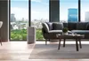 A modern living room with a large window overlooking a city skyline, featuring a sofa, coffee table, and an air purifier. Podgląd zdjęcia produktu
