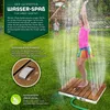 A young girl is playing in a garden sprinkler system, standing on a wooden shower platform. Náhled obrázku produktu