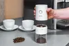 A hand holding the top part of a white coffee grinder over the bottom part, with coffee beans and cups in the foreground. Podgląd zdjęcia produktu