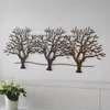 A metal wall decoration of three trees on a white background, with a plant in a pot in the foreground. Produktbild-Vorschau