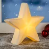 A lit-up star decoration sits on a surface covered in artificial snow, with red baubles in the background and a blurred snowy window scene behind it. Produktbild-Vorschau