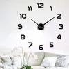 A wall clock with large, black, adhesive numbers and clock hands is mounted on a white wall above a sofa. Náhled obrázku produktu