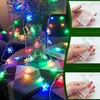 A string of colorful snowflake-shaped LED lights is draped around a vase of flowers and a book, with inset images showing how to insert batteries. Produktbild-Vorschau