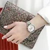 A person's arm wearing a watch is shown holding a glittery clutch bag. Produktbild-Vorschau