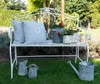 A white metal garden bench with two light gray cushions and potted plants, with watering cans and pitchers on the grass in front. Produktbild-Vorschau