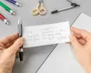 Hands holding a piece of paper with mathematical equations and a graph, with pens, scissors, and a compass on a gray surface. Produktbild-Vorschau