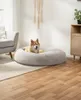 A dog is resting in a round, plush dog bed in a living room setting. Náhled obrázku produktu
