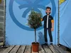 A potted olive tree stands next to a cardboard cutout of a man in front of a blue backdrop with a circular logo. Produktbild-Vorschau
