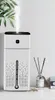 A white humidifier with a black top is shown on a white surface, with a plant and a diffuser in the background. Náhled obrázku produktu