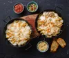 Two pans of baked pasta with melted cheese, served with small bowls of sauce and toppings, and toasted bread. Produktbild-Vorschau