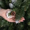 A hand holding a pine cone and a snowy branch against a backdrop of a Christmas tree. Voir les images du produit