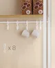 A close-up of a white metal rack with hooks and two white mugs hanging from it, against a beige wall. Náhled obrázku produktu