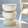 A cream-colored side table with a rounded, stacked design sits next to a chair in a room with concrete flooring and a gray wall. Produktbild-Vorschau