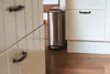 A stainless steel trash can with a pedal is placed on a wooden floor next to white kitchen cabinets. Produktbild-Vorschau
