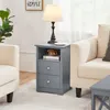 A gray nightstand with two drawers and an open shelf is placed next to a beige sofa in a living room setting. A lamp, a cup, and a decorative pumpkin are on the nightstand. Produktbild-Vorschau
