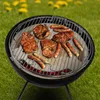 A close-up shot of a black kettle grill with various meats and sausages cooking on the grate, set against a blurred green grassy background with yellow flowers. Produktbild-Vorschau