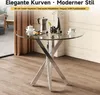 A round glass-top dining table with a modern metallic base, set in a room with wooden flooring and a curtained window. Náhľad obrázku produktu