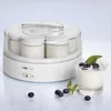 A white yogurt maker with several glass jars inside, placed next to a glass of yogurt with blueberries and mint. Produktbild-Vorschau