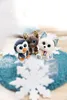 Three plush toys (a penguin, a reindeer, and a white dog/fox) are sitting on a blue knitted mat with white snowflake decorations. The background is blurred and light. Produktbild-Vorschau