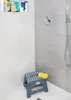 A small gray folding stool with a yellow sponge on top, placed in a shower stall with tiled walls. Náhled obrázku produktu