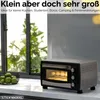 A small toaster oven with food inside, sitting on a countertop next to a basket of green apples, with a window and greenery in the background. Produktbild-Vorschau