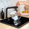 A person is filling a water filter pitcher under a black faucet in a kitchen sink. Náhľad obrázku produktu