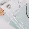 A close-up view of a gray roller blind with a metal eyelet and a decorative embroidered wave pattern. The blind is laid flat on a surface with some sliced oranges and a magazine in the background. Produktbild-Vorschau