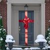 A decorated front door with a large red bow and ribbon, set against a brick house exterior with snow on the ground and bushes. Náhled obrázku produktu