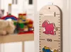 A wooden height chart with cartoon animal stickers is mounted on a wall. In the background, out of focus, are children's toys. Anteprima dell’immagine prodotto