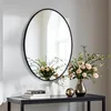 A round mirror with a black frame hangs on a wall above a console table. The table holds a vase of flowers, two decorative spheres, and a stack of books. Podgląd zdjęcia produktu