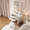 A white vanity table with drawers and a lighted mirror, a white stool, and a bed with a tufted headboard in a bedroom. Anteprima dell’immagine prodotto