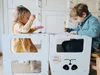 Two children are using a white learning tower in a kitchen. Anteprima dell’immagine prodotto