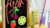 A chalkboard sign with drawings of fruit and the word "fresh" written on it, next to a glass of orange juice with a straw. Produktbild-Vorschau