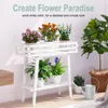 A white wooden plant stand with several potted plants on it, placed indoors with a large potted plant and a watering can nearby. Produktbild-Vorschau
