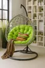A hanging egg chair with a green cushion and a brown throw blanket, placed in a room with a window and a bookshelf. Voir les images du produit