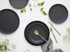 A flat lay of black plates and gold cutlery on a white background, with some greenery and white flowers. Podgląd zdjęcia produktu