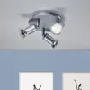 A silver-colored ceiling light fixture with three adjustable spotlights is mounted on a white ceiling. Below the light fixture, a blue wall is visible with two framed pieces of art. Voir les images du produit