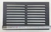 A black metal grate with horizontal slots is placed on a white surface, with a measuring tape along the bottom edge. Voir les images du produit