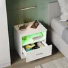 A white nightstand with a drawer open, showing items inside, next to a bed. The nightstand has LED lighting. Produktbild-Vorschau